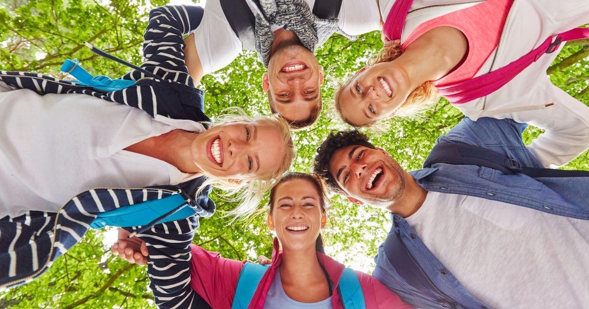 A diverse group of adults standing in a circle outdoors, smiling and connecting joyfully, with sunlight and green leaves in the background.
